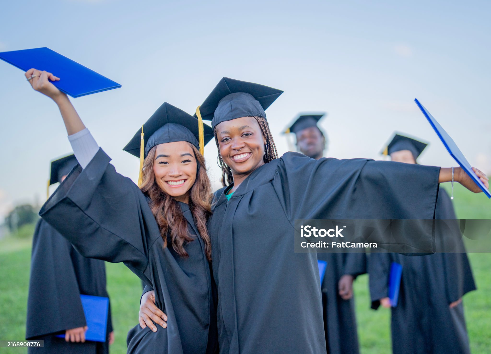 Two female university graduates pose together outside for a portrait as they celebrate the completing of their education. They are dressed in formal black gowns and have caps on as they hold their diplomas.