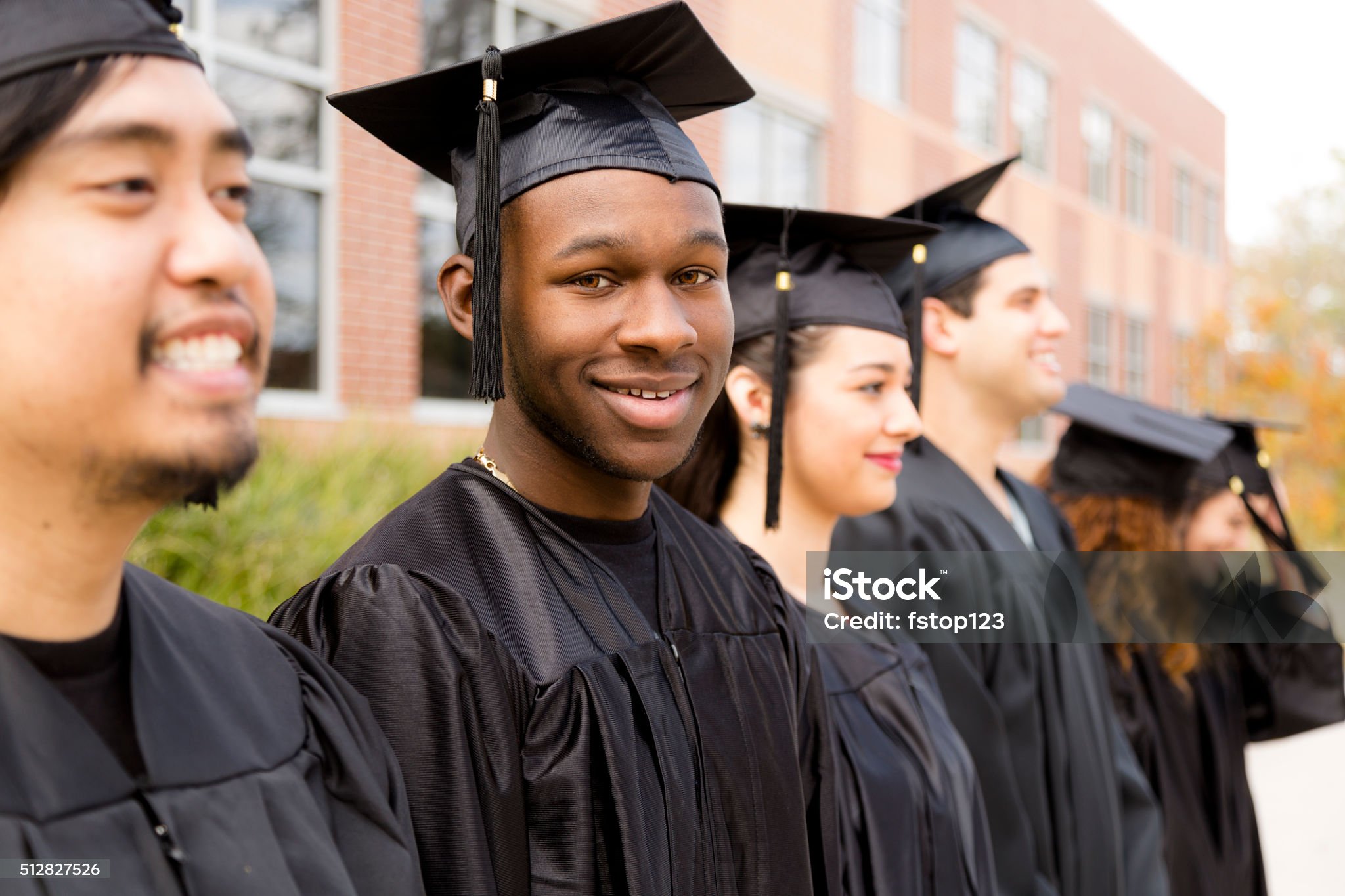Six, multi-ethnic friend graduates excitedly wait for their name to be called during graduation ceremony. African descent male looks back at camera. School building background.